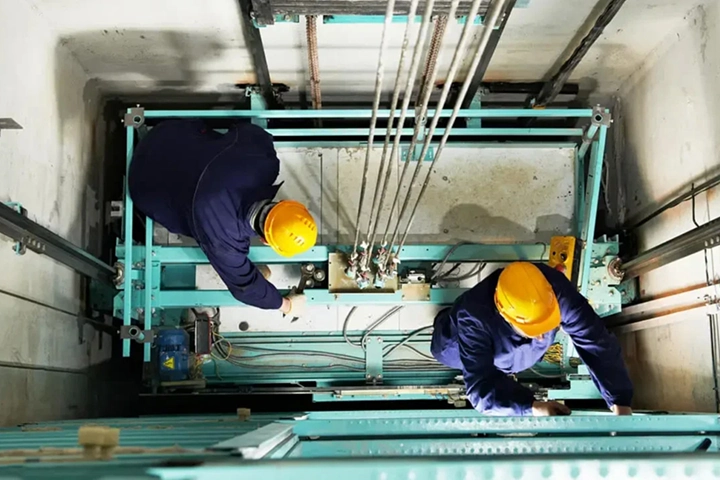 Home 18 Two expert technicians performing a new elevator installation service in Chennai, seen from above as they work on the machinery on top of the lift car.